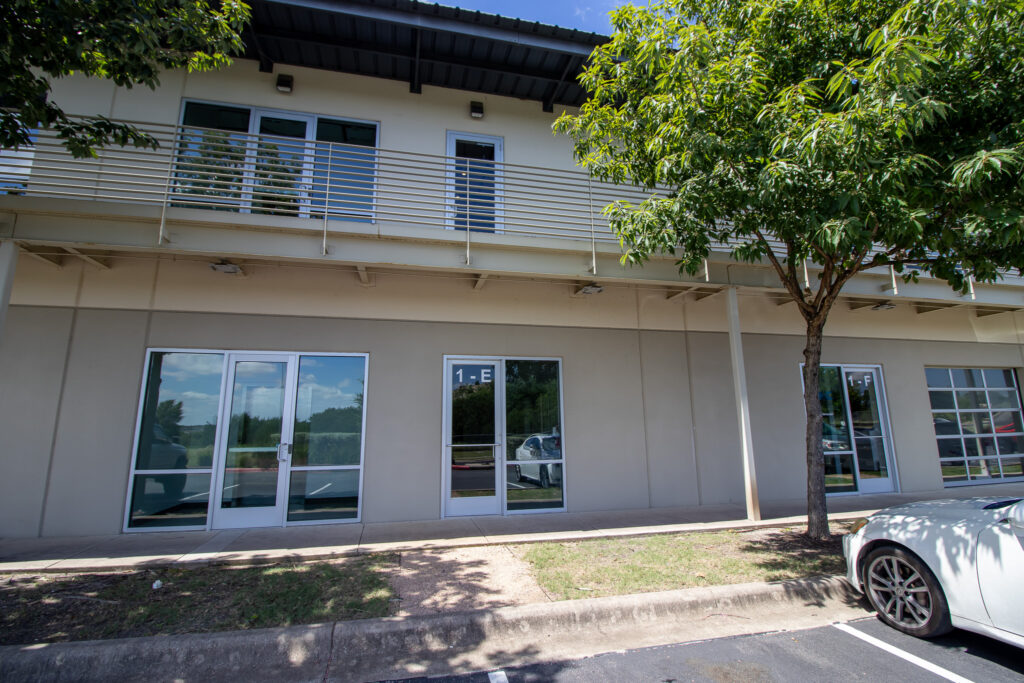 Exterior entrance of Suite E2 at 1205 Sheldon Cove, Austin, featuring glass double doors, parking, and tree-lined frontage.